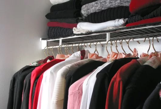 Various styles of shirt hanging in a closet with sweaters stacked above them on a shelf