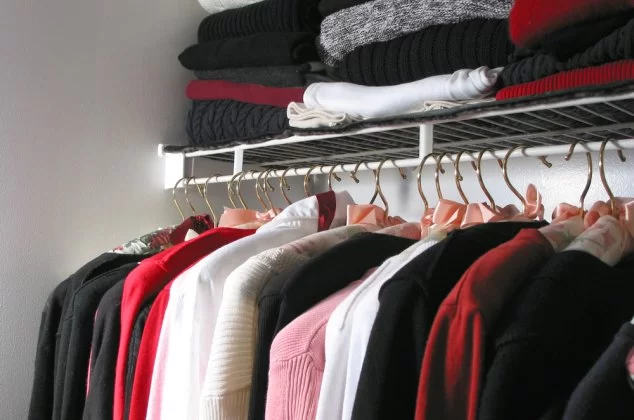 Various styles of shirt hanging in a closet with sweaters stacked above them on a shelf