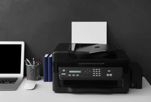 Black printer on a desk next to pens, a computer, and other office items