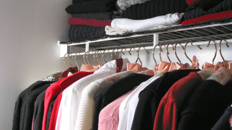 Various styles of shirt hanging in a closet with sweaters stacked above them on a shelf