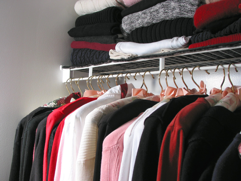 Various styles of shirt hanging in a closet with sweaters stacked above them on a shelf