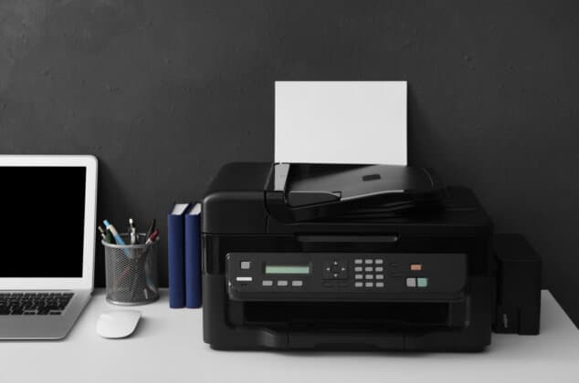 Black printer on a desk next to pens, a computer, and other office items