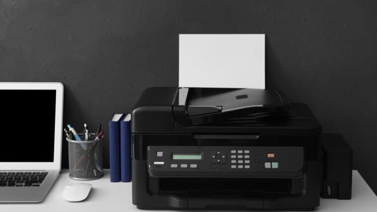 Black printer on a desk next to pens, a computer, and other office items